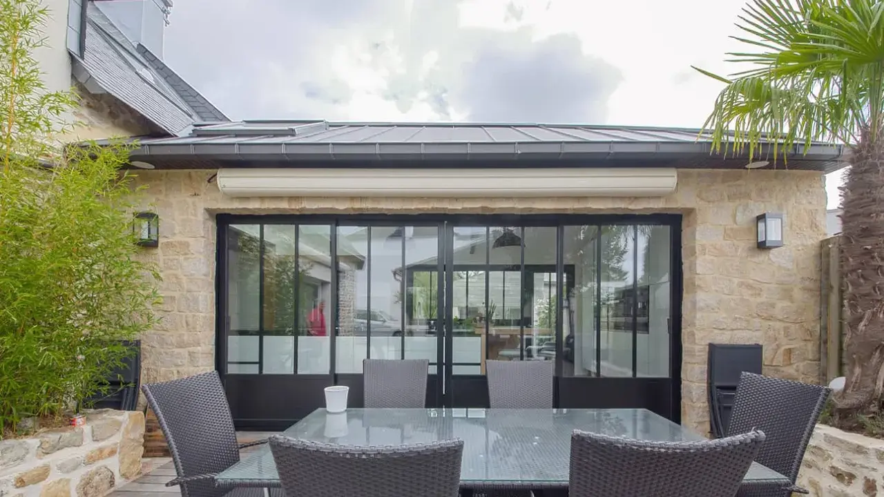 Terrasse avec mobilier autour d'une extension maison style atelier aux grandes verrières noires. Bambou et palmier apportent une touche végétale.