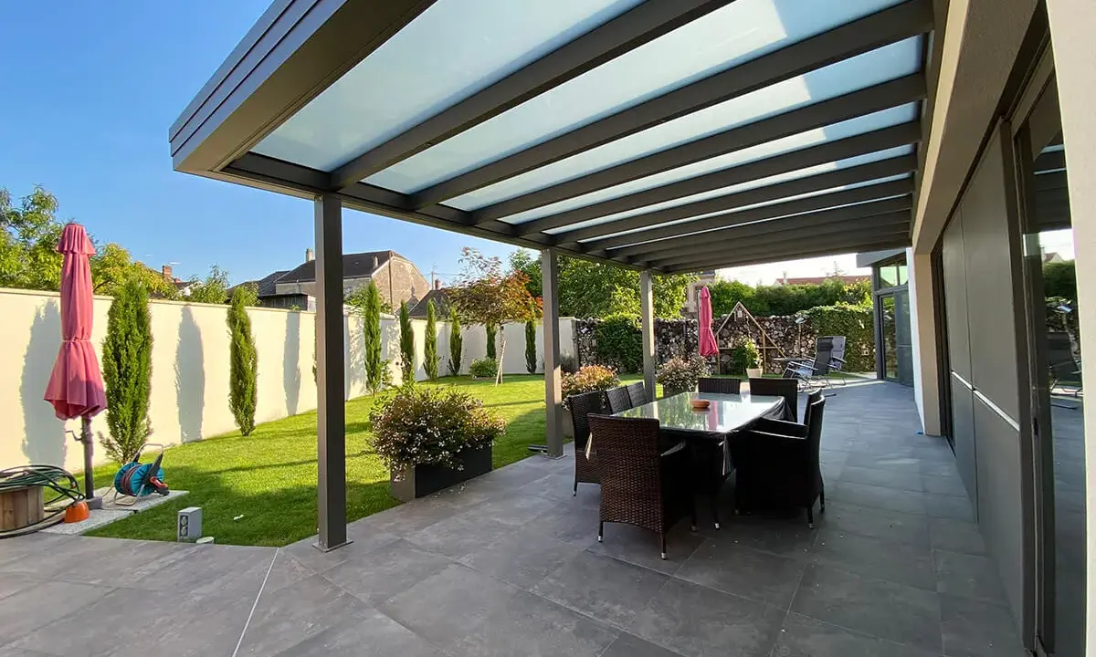 Une belle pergola moderne avec un toit vitré pour couvrir l'espace repas extérieur. Le jardin est bien entretenu, avec une pelouse verte et des cyprès élancés.