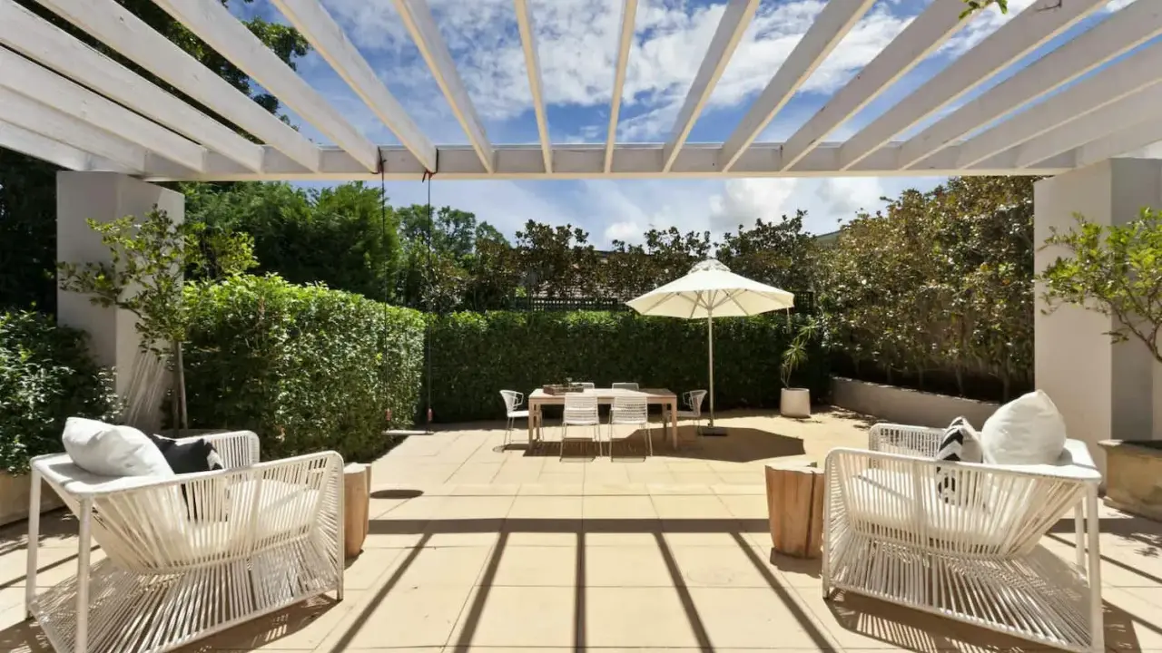 Terrasse couverte avec pergola blanche, mobilier de jardin blanc, table et parasol. Un espace extérieur aménagé, peut-être pour fermer une terrasse couverte sans autorisation.