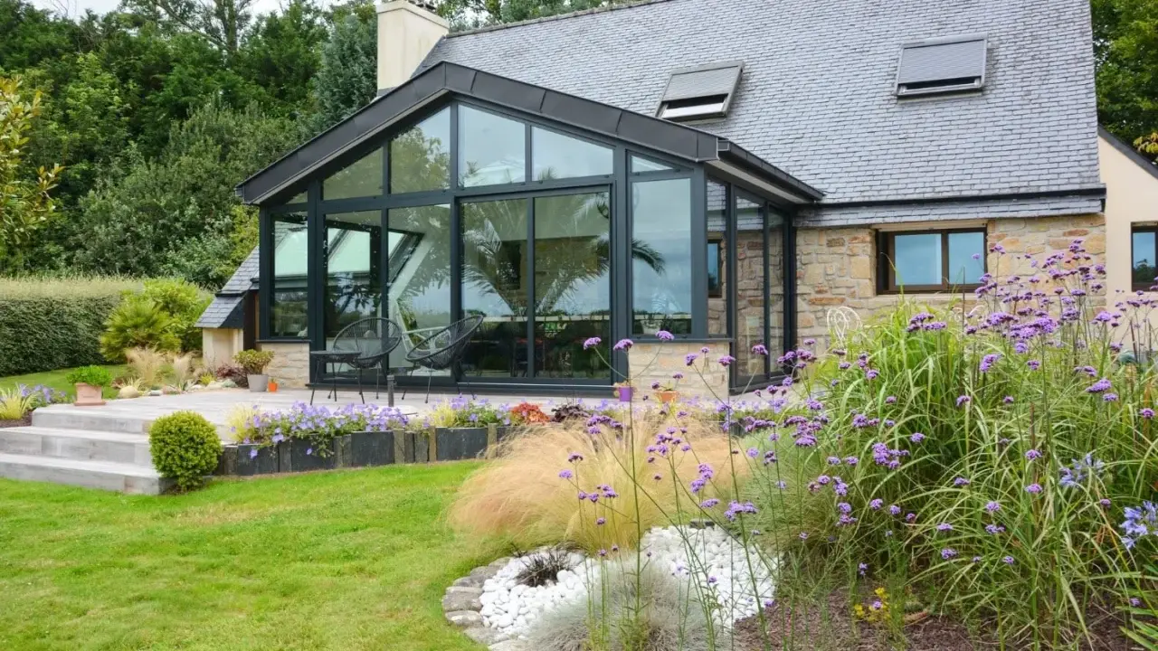 Maison avec extension toit vitrée, terrasse en bois et jardin fleuri.