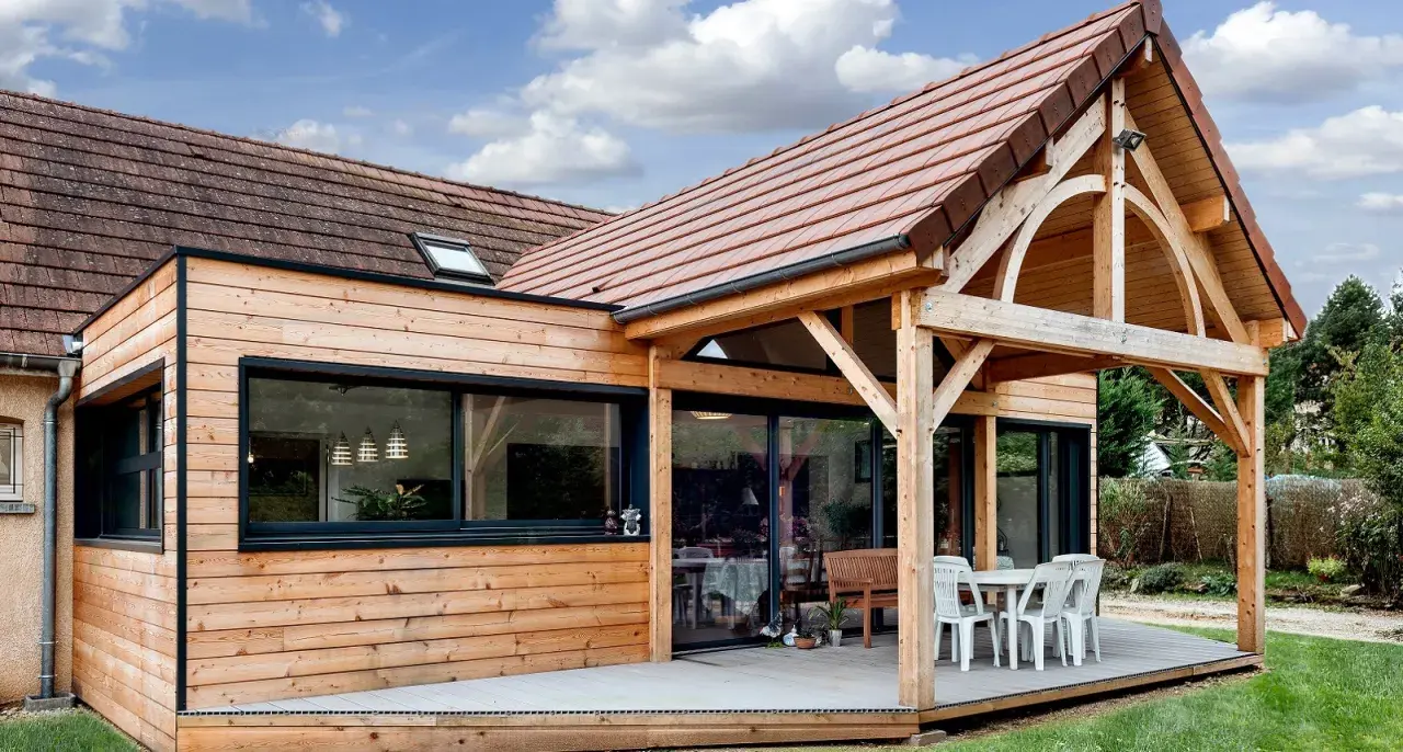 Extension de maison en bois avec terrasse sur dalle béton. Une table et des chaises sont installées sous un auvent.