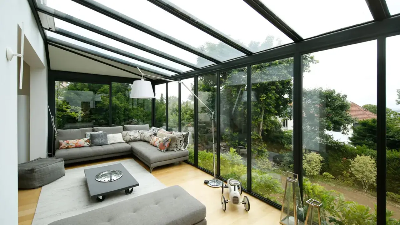Salon lumineux avec canapé d'angle gris, table basse et vue sur jardin luxuriant. Idéal pour agrandir veranda et profiter de la nature.
