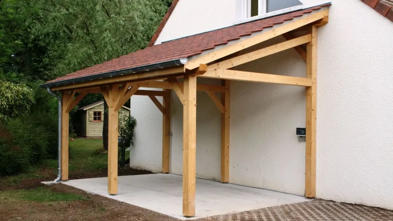 Un abri de voiture en bois avec une pente légère, adossé à une maison blanche.