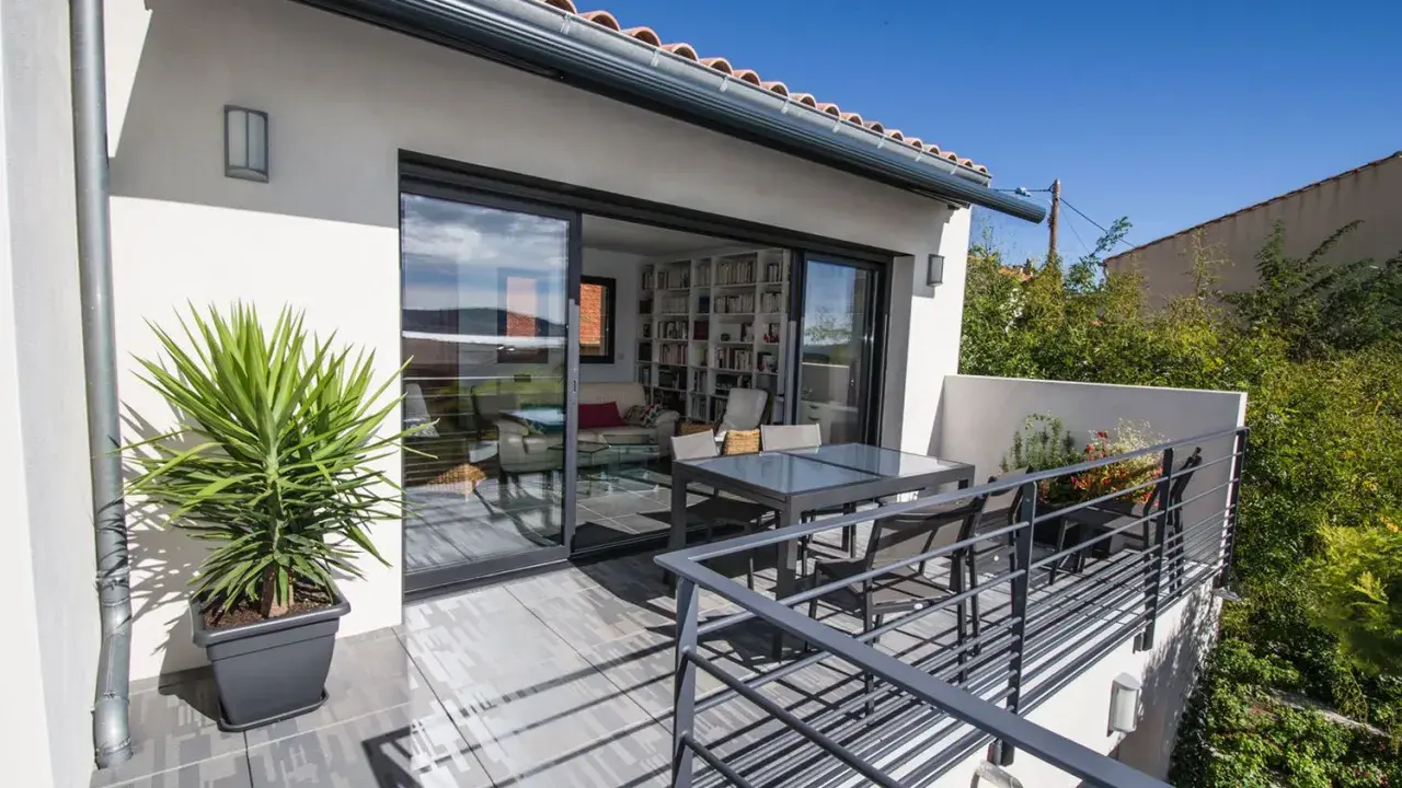 Terrasse sur le toit accessible, avec table et chaises, et une grande baie vitrée donnant sur un salon rempli de livres.