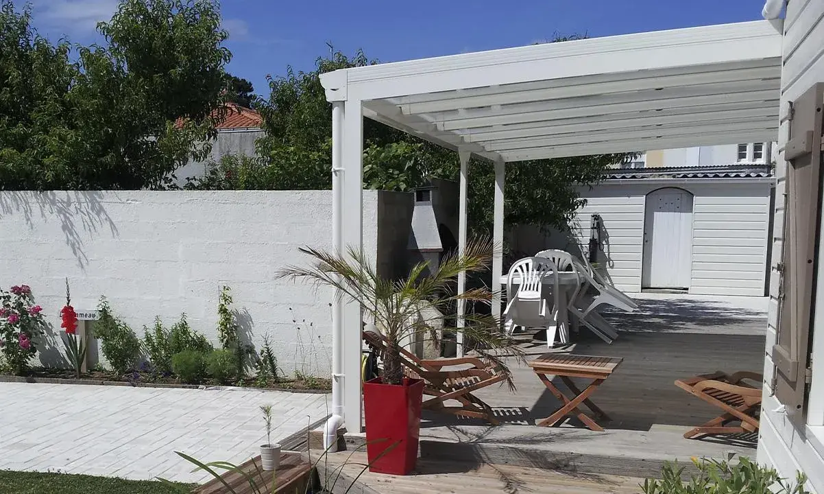 Terrasse en bois sous un préau semi-fermé, avec chaises empilées, une table, un palmier en pot et un barbecue.