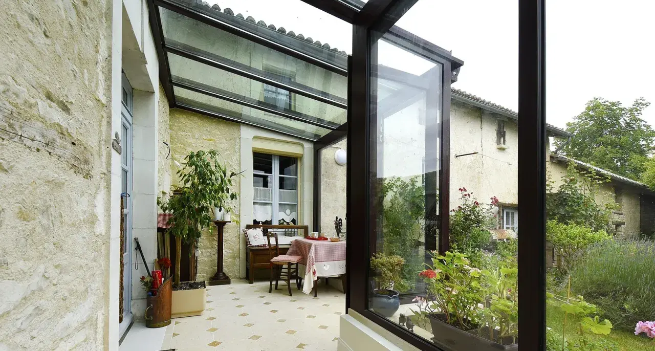 Une veranda avec toiture tuile, aménagée avec une table et des plantes, s'ouvre sur un jardin verdoyant.