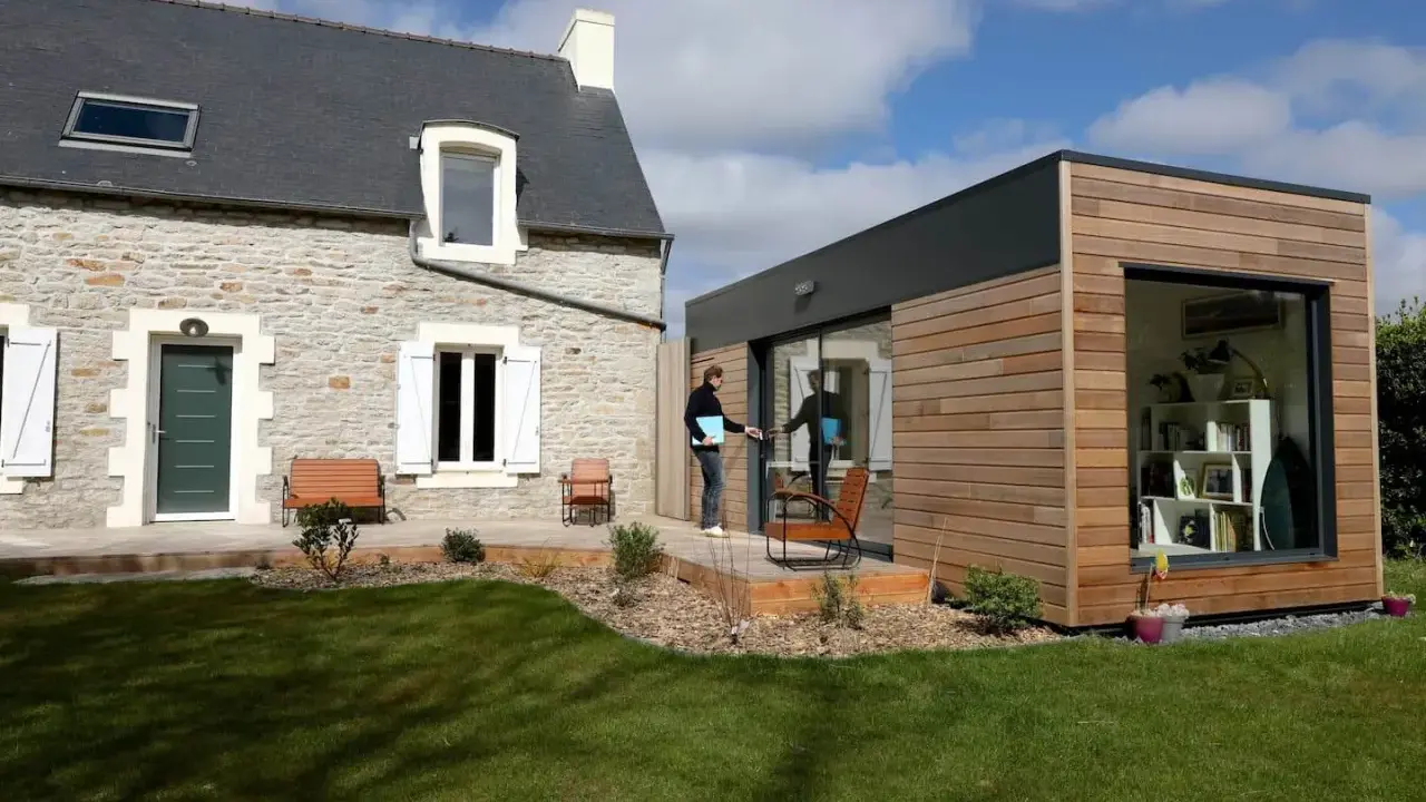 Une maison en pierre avec une extension moderne en bois. Un homme sort de l'extension, prêt à profiter du jardin.