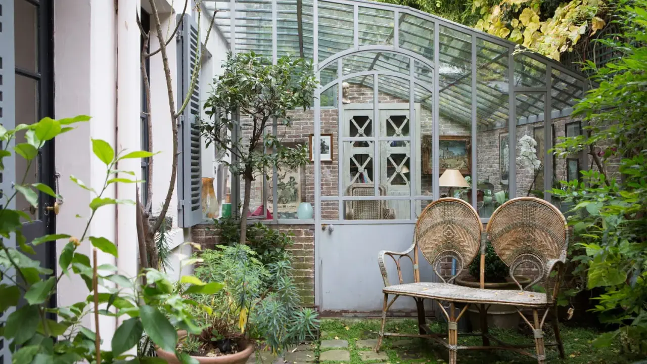 Une charmante veranda en fer forgé abrite un banc en rotin et des plantes luxuriantes, invitant à la détente dans ce jardin secret.
