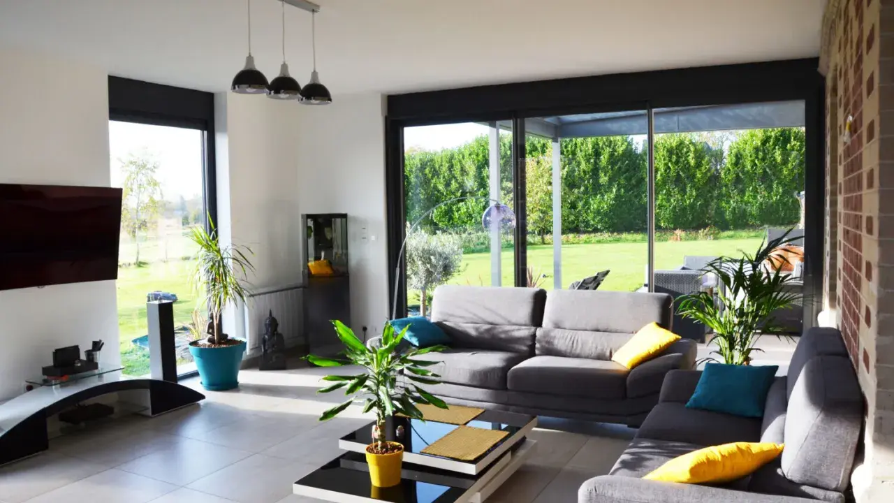 Salon lumineux avec canapés gris, table basse moderne et grandes baies vitrées donnant sur un jardin verdoyant. Une annexe maison parfaite pour se détendre.