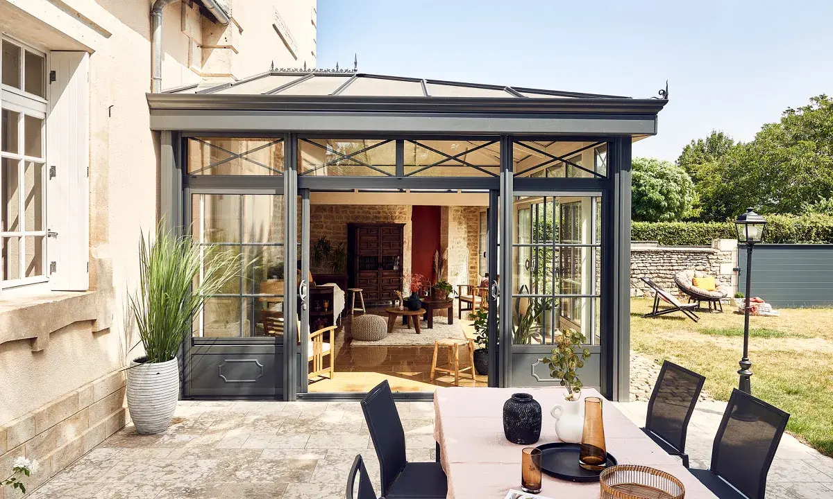 Une véranda moderne s'ouvre sur un jardin verdoyant. Une table dressée invite à profiter de l'extérieur, peut-être pour éviter la taxe terrasse.