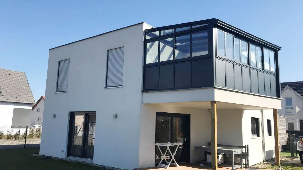 Maison moderne avec une veranda 2 niveaux vitrée, offrant une extension lumineuse. Terrasse en bois en contrebas.