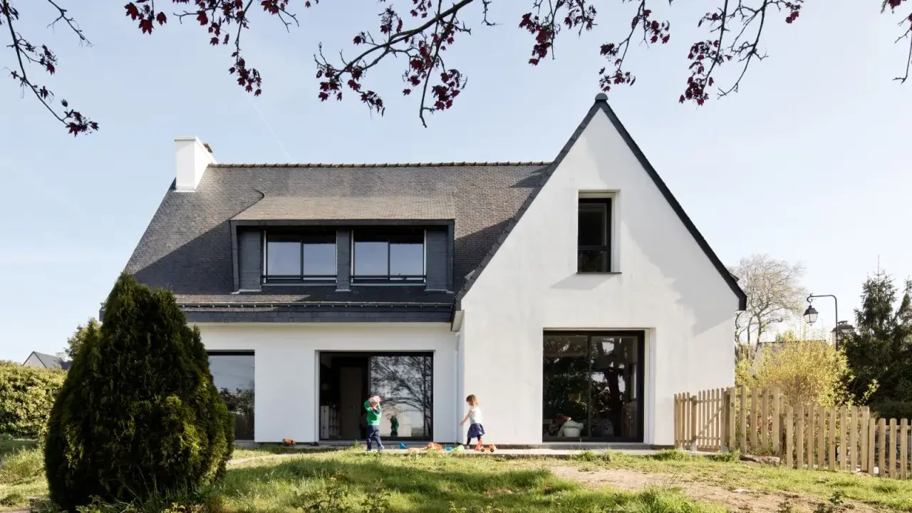Une extension maison néo-bretonne moderne avec de grandes baies vitrées. Deux enfants jouent dans le jardin verdoyant devant la maison.