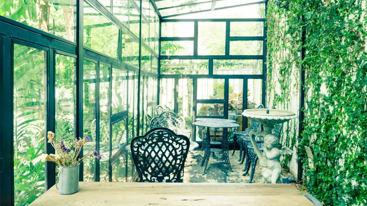 Un charmant jardin d'hiver verrière, avec des tables et chaises en fer forgé, une statue d'ange et une profusion de verdure.