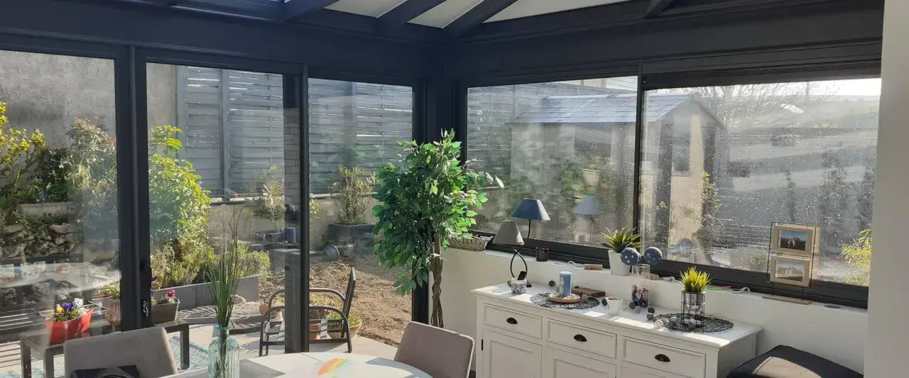 Intérieur lumineux d'une véranda triple vitrage, avec vue sur un jardin verdoyant et une commode blanche décorée.