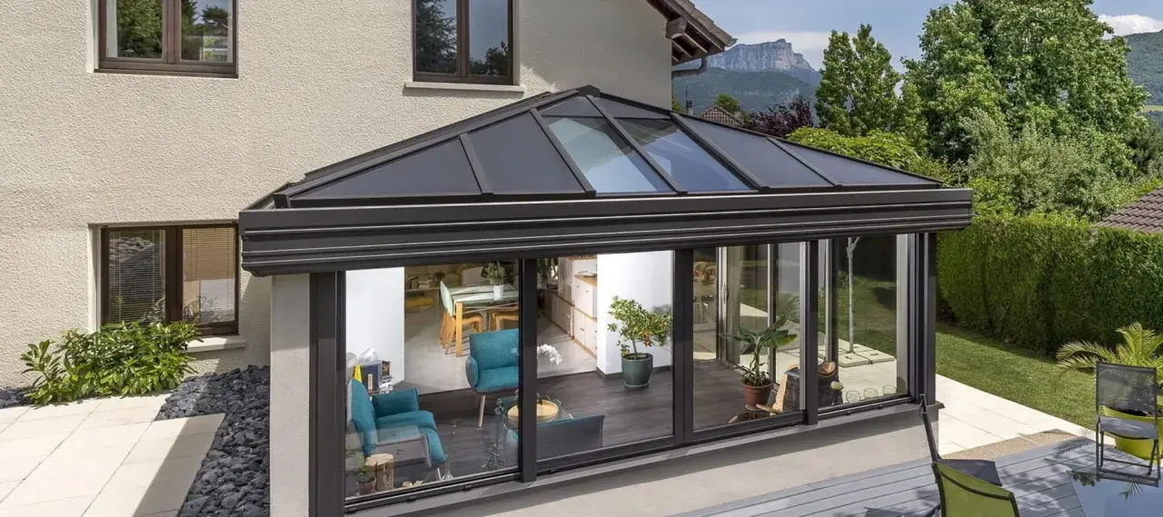 Une **pergola bioclimatique** moderne avec toit vitré, prolongeant une maison vers un jardin verdoyant. L'intérieur lumineux révèle un salon cosy et une salle à manger.