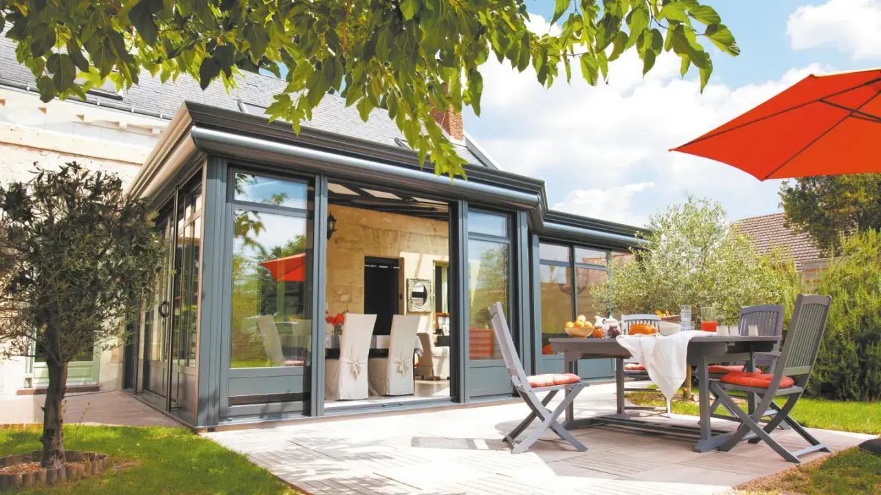 Une véranda moderne avec un surbot béton, une table et des chaises sur une terrasse en bois, sous un parasol orange vif.