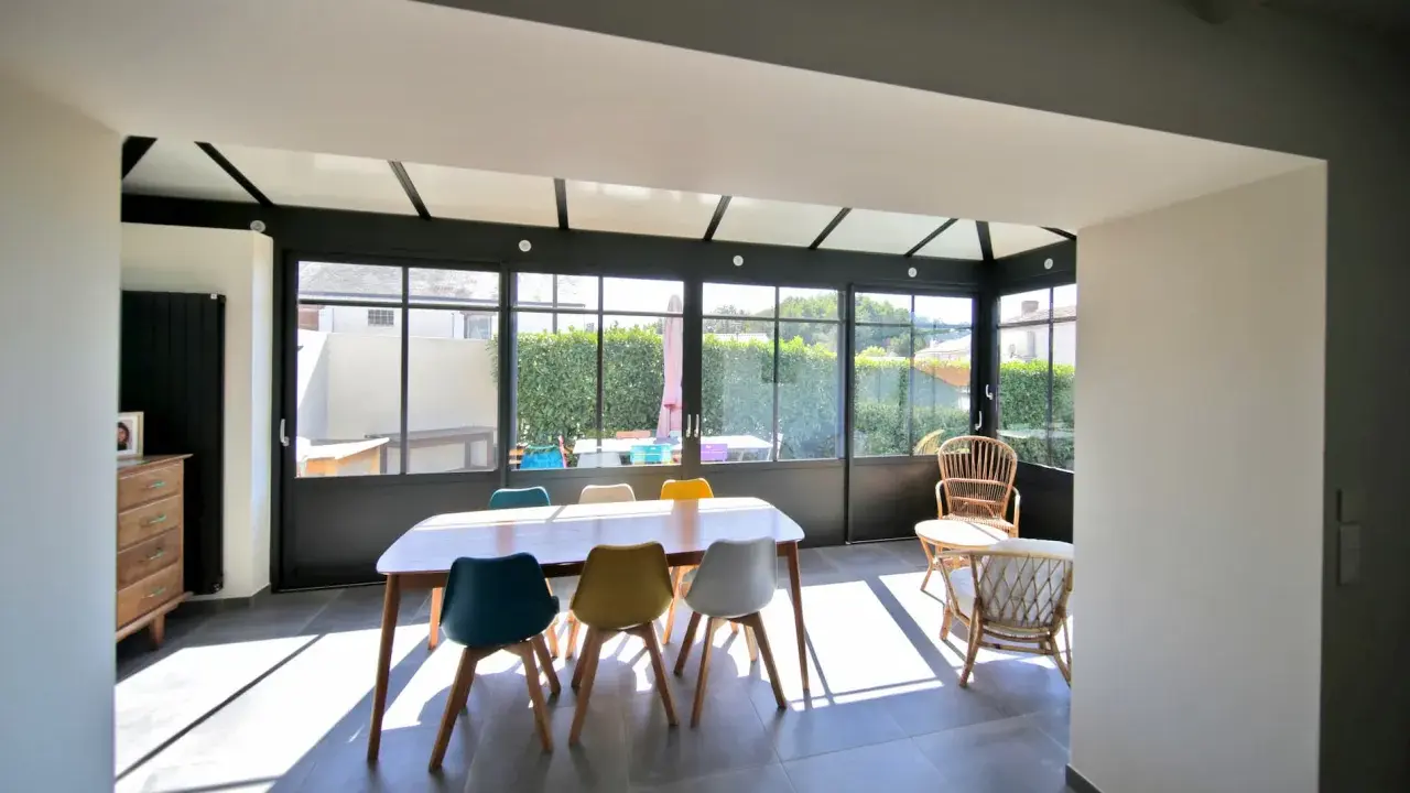 Salle à manger lumineuse dans une véranda chauffée, avec une table en bois et des chaises colorées. Un espace de vie agréable.