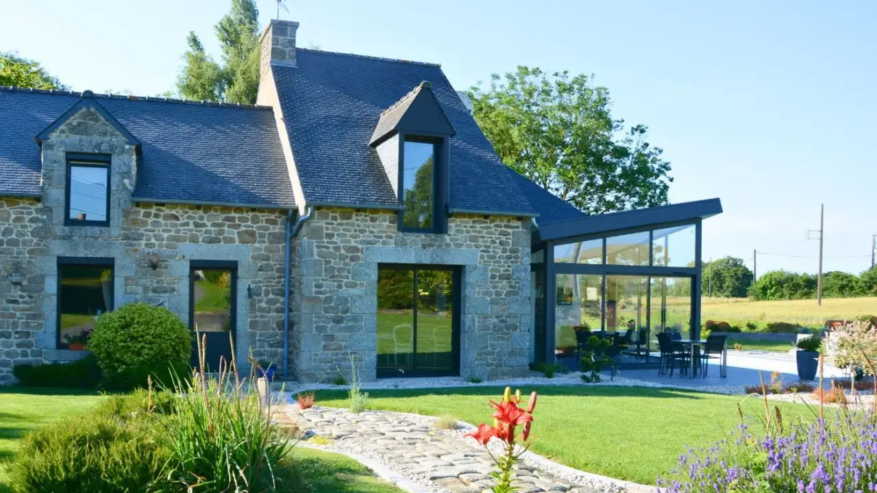 Maison en pierre avec une extension longere vitrée moderne. Un chemin pavé mène à la terrasse où une table est dressée.