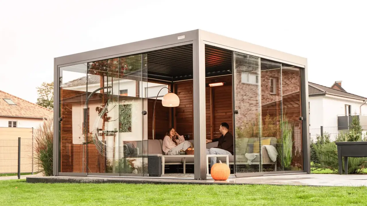 Un couple se détend dans une pergola moderne avec parois vitrées. La fixation pergola est discrète, laissant place à un espace cosy avec chauffage d'appoint.