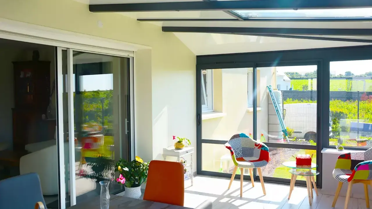 Intérieur lumineux d'une véranda avec un grand vitrage offrant une vue sur un jardin verdoyant. Des chaises colorées invitent à la détente.