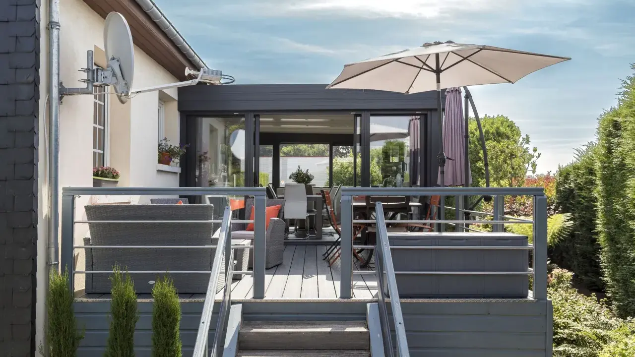 Terrasse avec mobilier de jardin et une véranda bois et alu. Parasol ouvert, escalier menant à la terrasse.