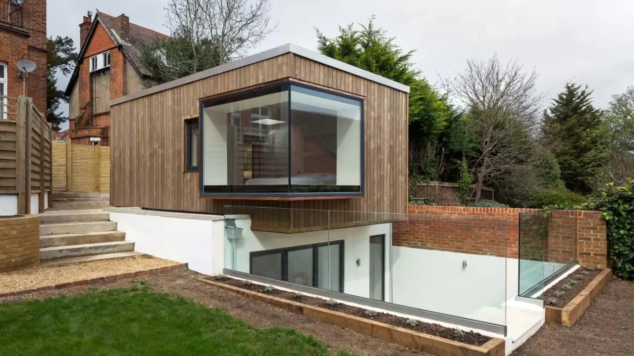 Une extension de maison moderne en bois avec une grande fenêtre en saillie. L'agrandissement maison s'intègre à un jardin en pente avec des murs de briques et des garde-corps en verre.