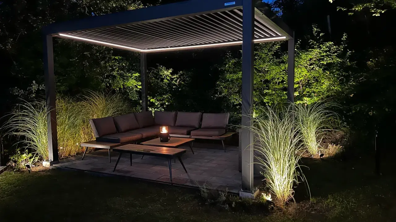 Une **pergola au milieu du jardin** illuminée la nuit, avec un salon de jardin confortable et des herbes hautes éclairées.