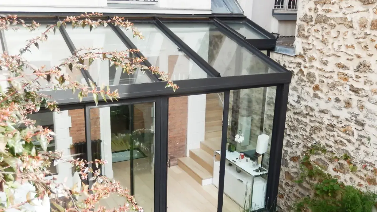 Une pergola transformée en véranda moderne, avec une structure en verre et métal, s'intégrant à une façade en pierre et brique.