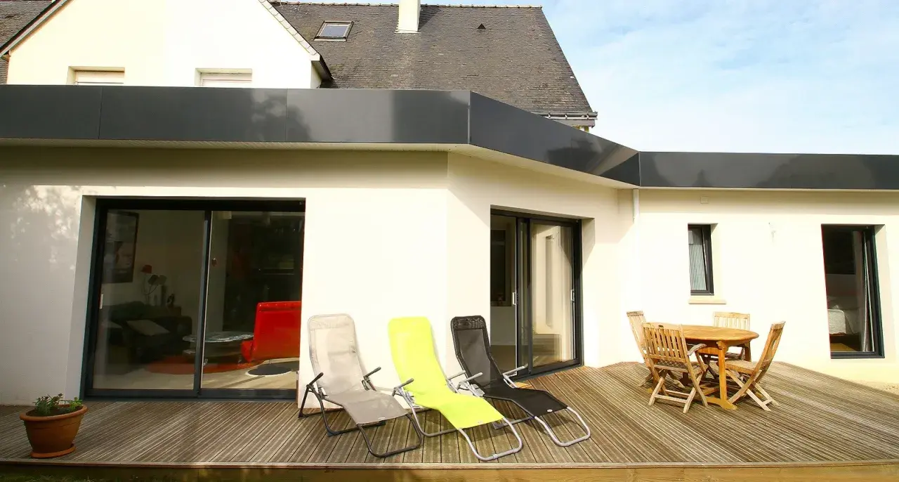 Extension de toit plat en bac acier, avec terrasse en bois, chaises longues et table de jardin.
