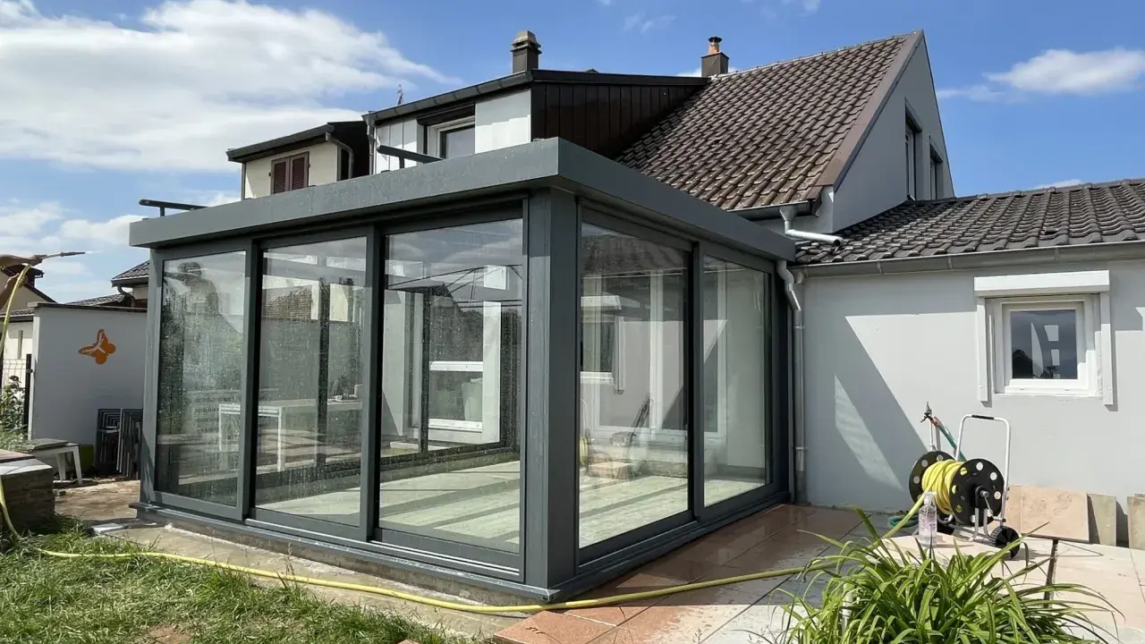 Une véranda moderne, conçue par un architecte, prolonge une maison. Elle est en verre et métal gris, avec un toit plat. Le jardin est verdoyant.