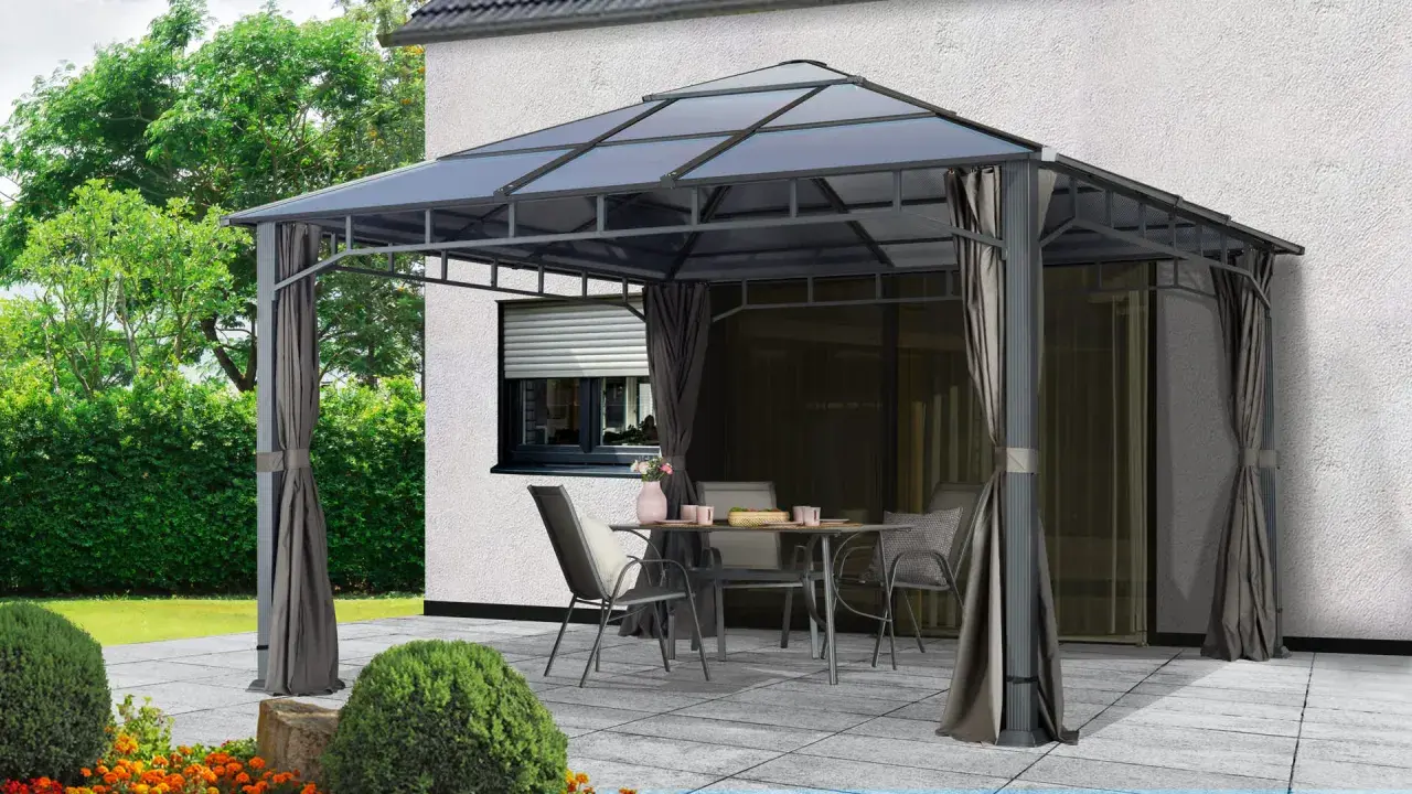 Une tonnelle def. avec table et chaises sur une terrasse pavée, devant une maison blanche et un jardin verdoyant.