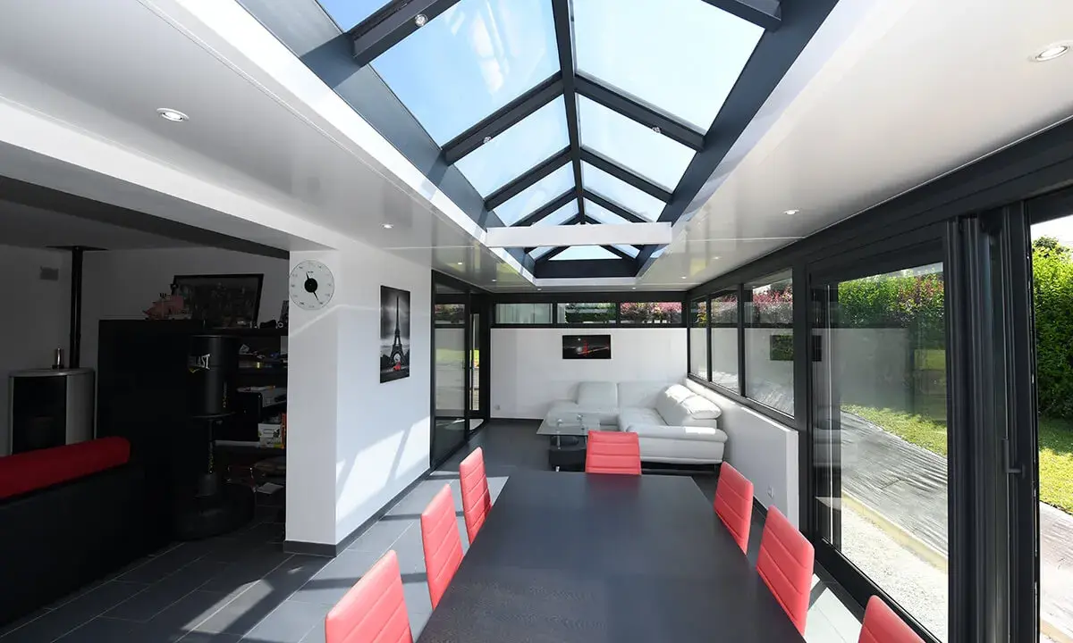 Une extension veranda salle à manger moderne avec un toit vitré, une table noire et des chaises rouges, un canapé blanc et une vue sur le jardin.