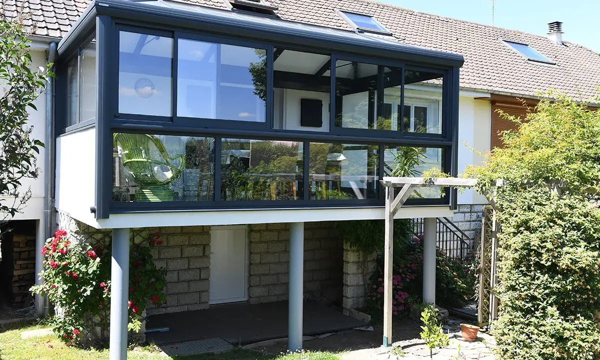 Moderne veranda adossée au mur, avec une chaise verte visible à travers les grandes baies vitrées. Le soleil brille sur le jardin verdoyant.