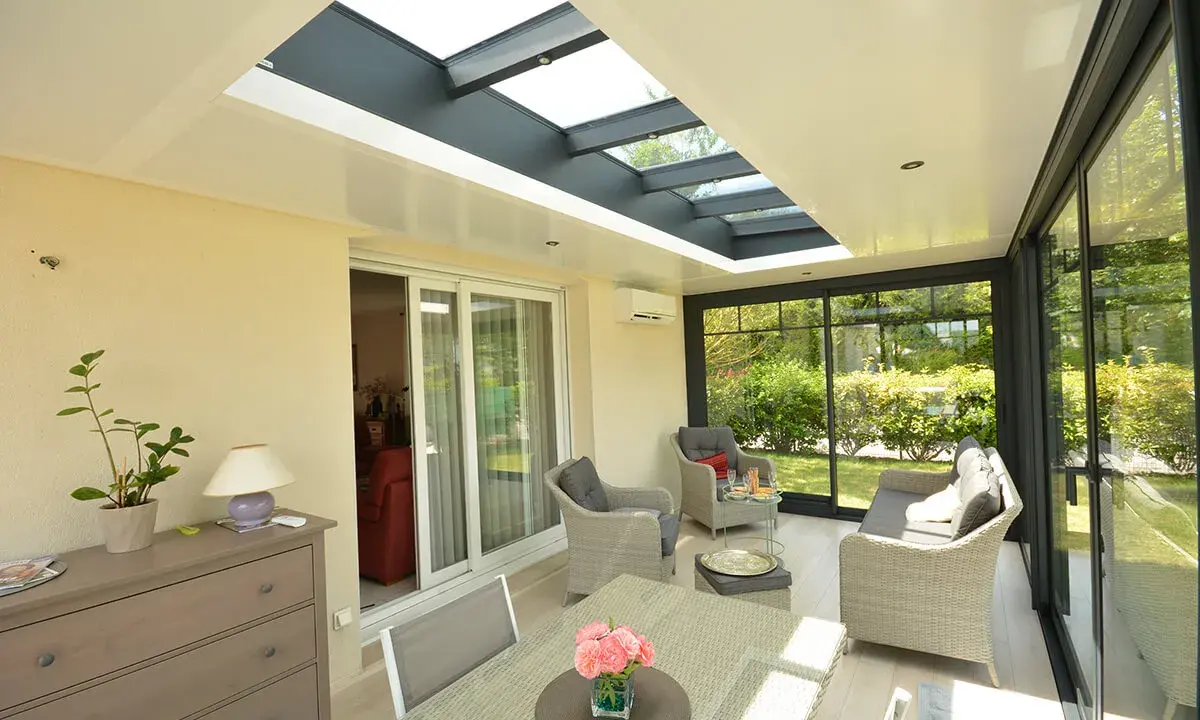 Une **veranda qui s'ouvre entièrement** sur un jardin verdoyant. Mobilier confortable, table et chaises pour profiter du soleil.