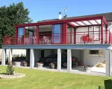 Une maison avec une veranda terrasse rouge vif, offrant une vue sur un jardin verdoyant et une voiture garée sous le balcon.