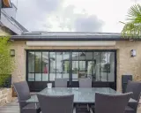 Terrasse avec mobilier autour d'une extension maison style atelier aux grandes verrières noires. Bambou et palmier apportent une touche végétale.