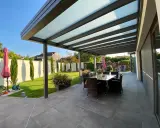 Une belle pergola moderne avec un toit vitré pour couvrir l'espace repas extérieur. Le jardin est bien entretenu, avec une pelouse verte et des cyprès élancés.