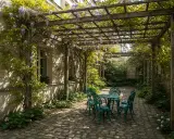 Un charmant modèle de pergola pour terrasse, envahi par la glycine, abrite une table et quatre chaises en fer forgé.