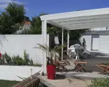 Terrasse en bois sous un préau semi-fermé, avec chaises empilées, une table, un palmier en pot et un barbecue.