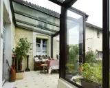 Une veranda avec toiture tuile, aménagée avec une table et des plantes, s'ouvre sur un jardin verdoyant.