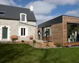 Une maison en pierre avec une extension moderne en bois. Un homme sort de l'extension, prêt à profiter du jardin.