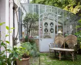 Une charmante veranda en fer forgé abrite un banc en rotin et des plantes luxuriantes, invitant à la détente dans ce jardin secret.