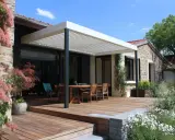 Construction d'une pergola moderne sur une terrasse en bois, avec une table et des chaises, devant une maison en pierre.