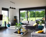 Salon lumineux avec canapés gris, table basse moderne et grandes baies vitrées donnant sur un jardin verdoyant. Une annexe maison parfaite pour se détendre.