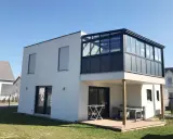 Maison moderne avec une veranda 2 niveaux vitrée, offrant une extension lumineuse. Terrasse en bois en contrebas.