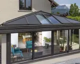 Une **pergola bioclimatique** moderne avec toit vitré, prolongeant une maison vers un jardin verdoyant. L'intérieur lumineux révèle un salon cosy et une salle à manger.