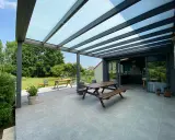 Terrasse avec table en bois sous une pergola bioclimatique. La hauteur de la structure permet de profiter du jardin verdoyant.