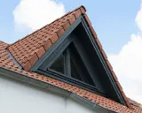 Un puit de lumière triangulaire s'intègre dans un toit en tuiles rouges, sous un ciel bleu et nuageux.