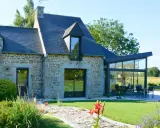 Maison en pierre avec une extension longere vitrée moderne. Un chemin pavé mène à la terrasse où une table est dressée.