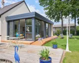Extension toit plat sur maison ancienne, avec terrasse en bois, mobilier de jardin et vue sur un jardin verdoyant.